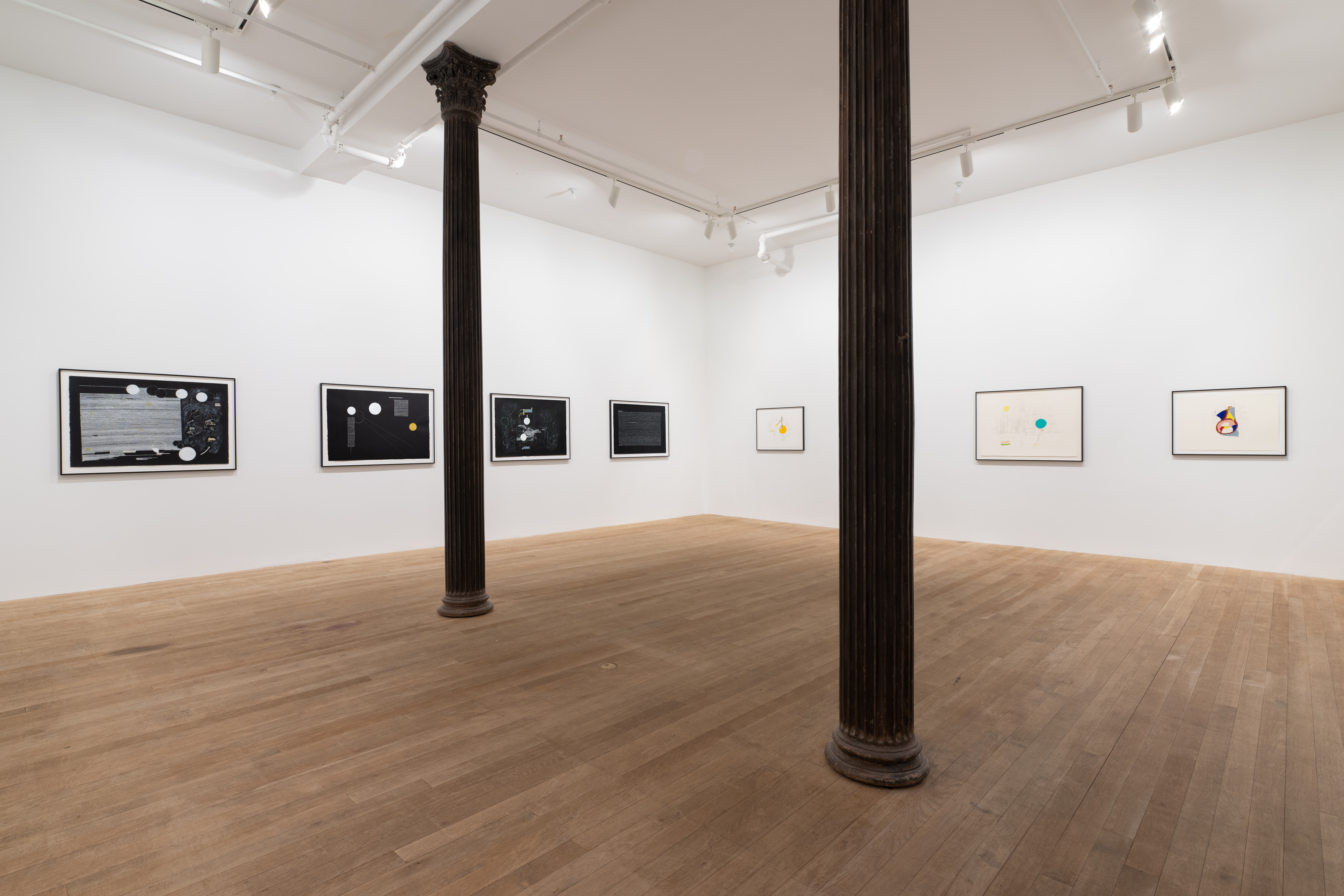 A color image of a gallery space with two columns running across the room. On the left wall, four framed abstract drawings on black paper are displayed in a horizontal line. On the right wall, three framed abstract drawings on white paper are shown in a horizontal line.