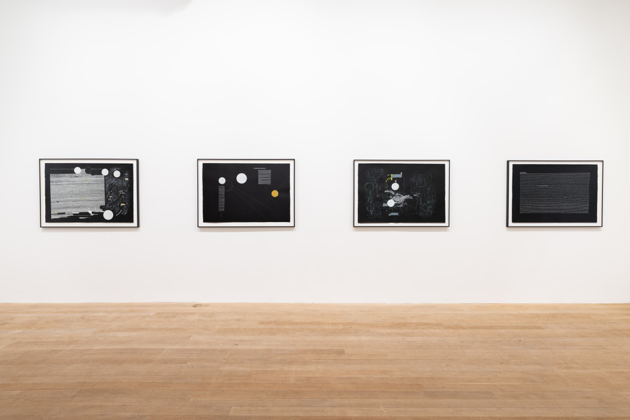 A color image of four framed works displayed in a horizontal line on a white wall. The drawings, made on black paper, depict abstract compositions made up of white lines, arrows, circles, and illegible textlines.