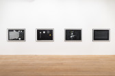 A color image of four framed works displayed in a horizontal line on a white wall. The drawings, made on black paper, depict abstract compositions made up of white lines, arrows, circles, and illegible textlines.