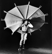 A person on roller skates and knee pads with a large, fabric umbrella held on his back.