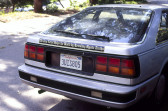 A black bumper sticker with white text on the back of a silver car reads, 