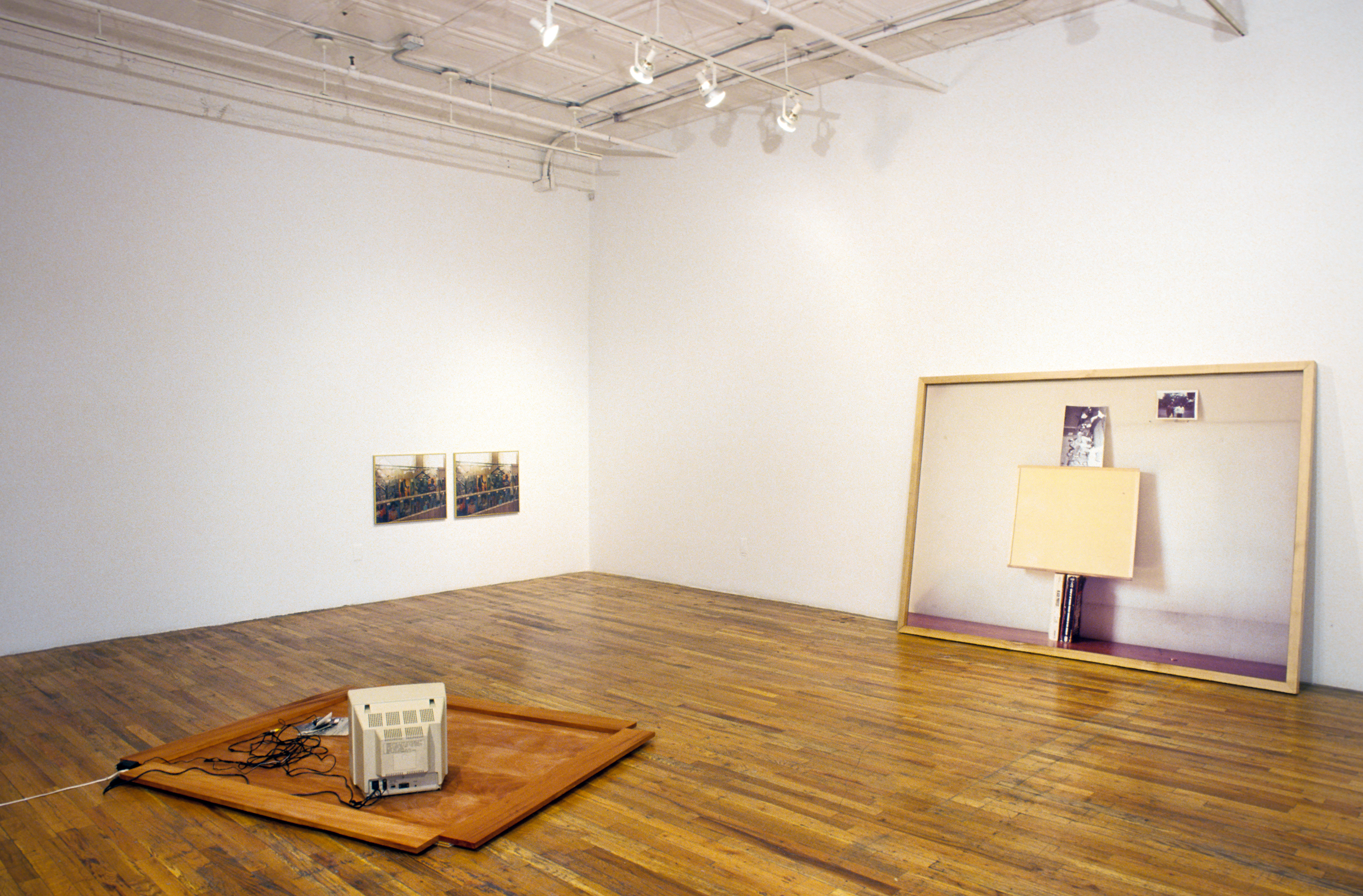 A gray television sits on a rectangular wooden board. There is a mass of wires extending from the back of it snaking towards a pile of nearby photographs. On one white wall opposite, there are two identical images. Leaning on the other white wall is a framed photograph of two books, a wooden rectangle, and a photograph stacked vertically on each other. There is a second photograph offset to the right.