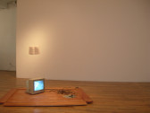 A gray television sits on a rectangular wooden board. There is a mass of wires next to it. In the background, a white paper with black text hangs on a white wall.