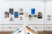 A view of a book stand perpendicular to a wall in which staggered posters are hung. The book stand displays various documents and materials.