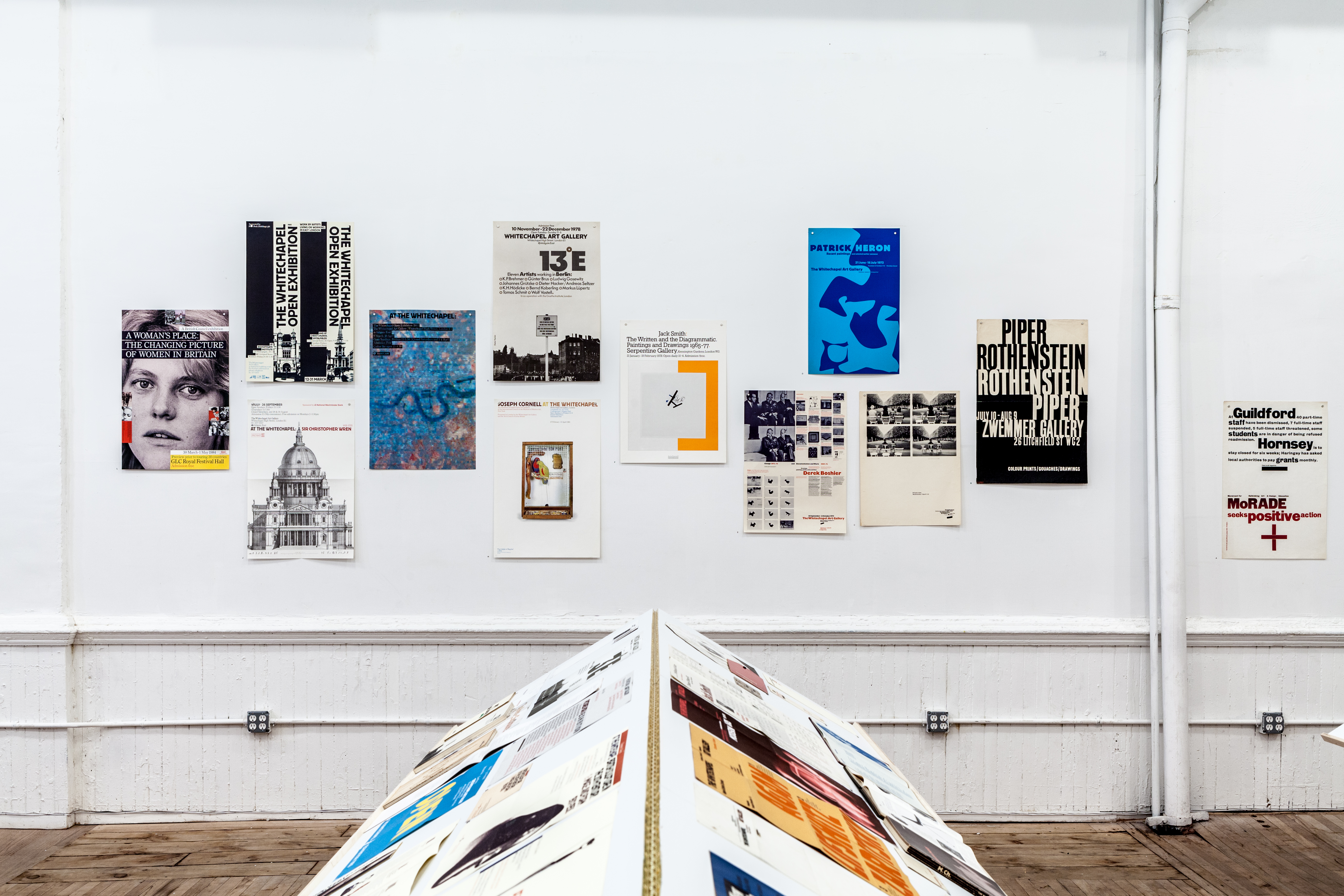 A view of a book stand perpendicular to a wall in which staggered posters are hung. The book stand displays various documents and materials.