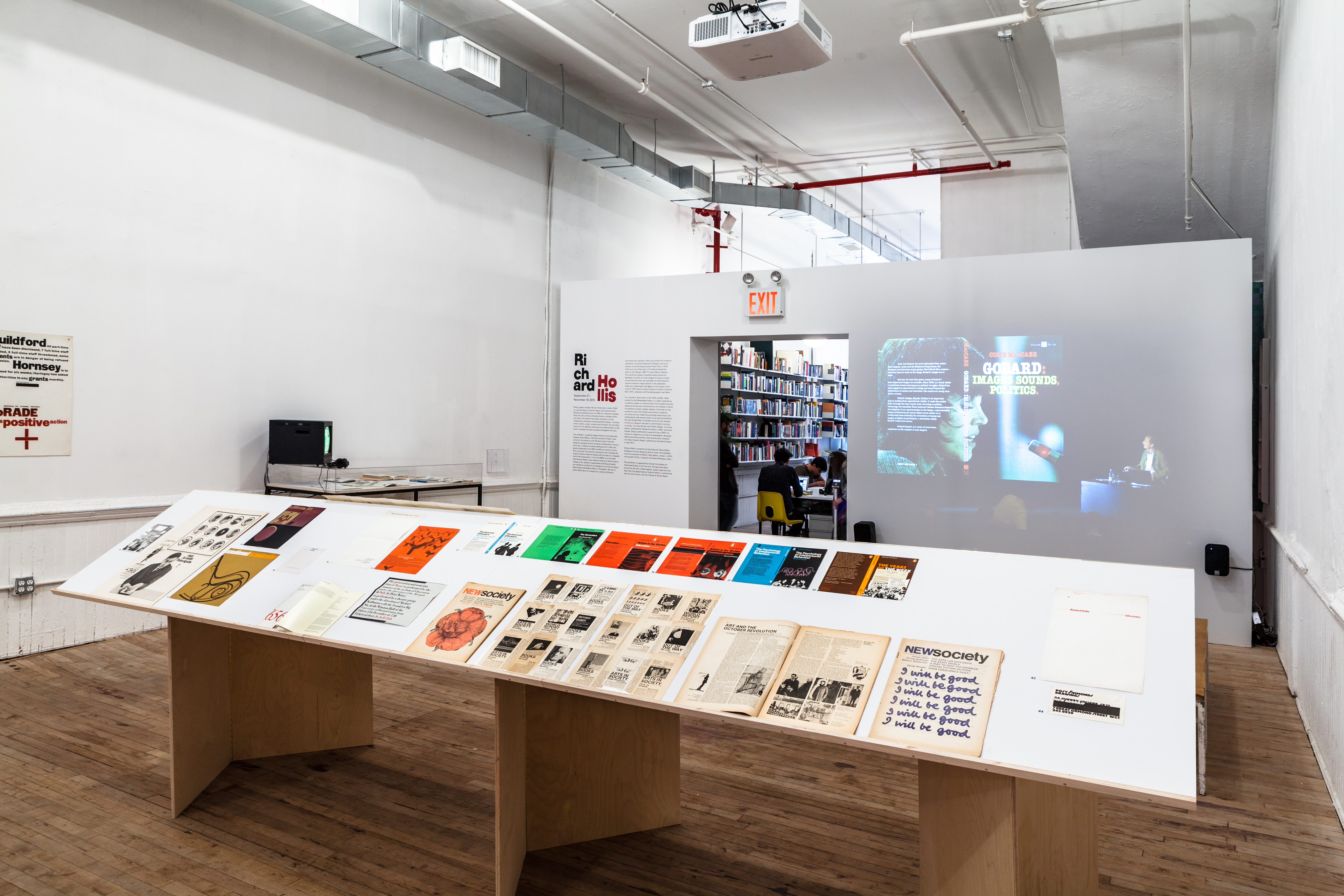 An angled view of the exhibition exit, on which the left of the doorway is text, on the right a projected image. Across the frame is a large bookstand displaying documents, books, and papers.
