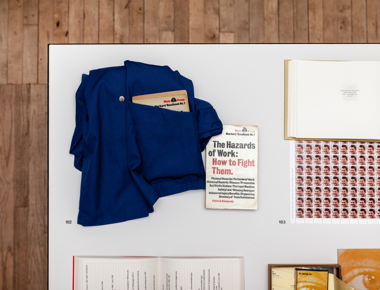 A blue shirt sits folded on a table with a book inside its pocket. Next to it, a book with text on the cover that reads, "The Hazards of Work: How to Fight Them."
