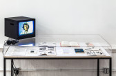 A small television on top of a vitrine shows John Berger looking at the viewer. In the vitrine, several books and documents are opened for display.
