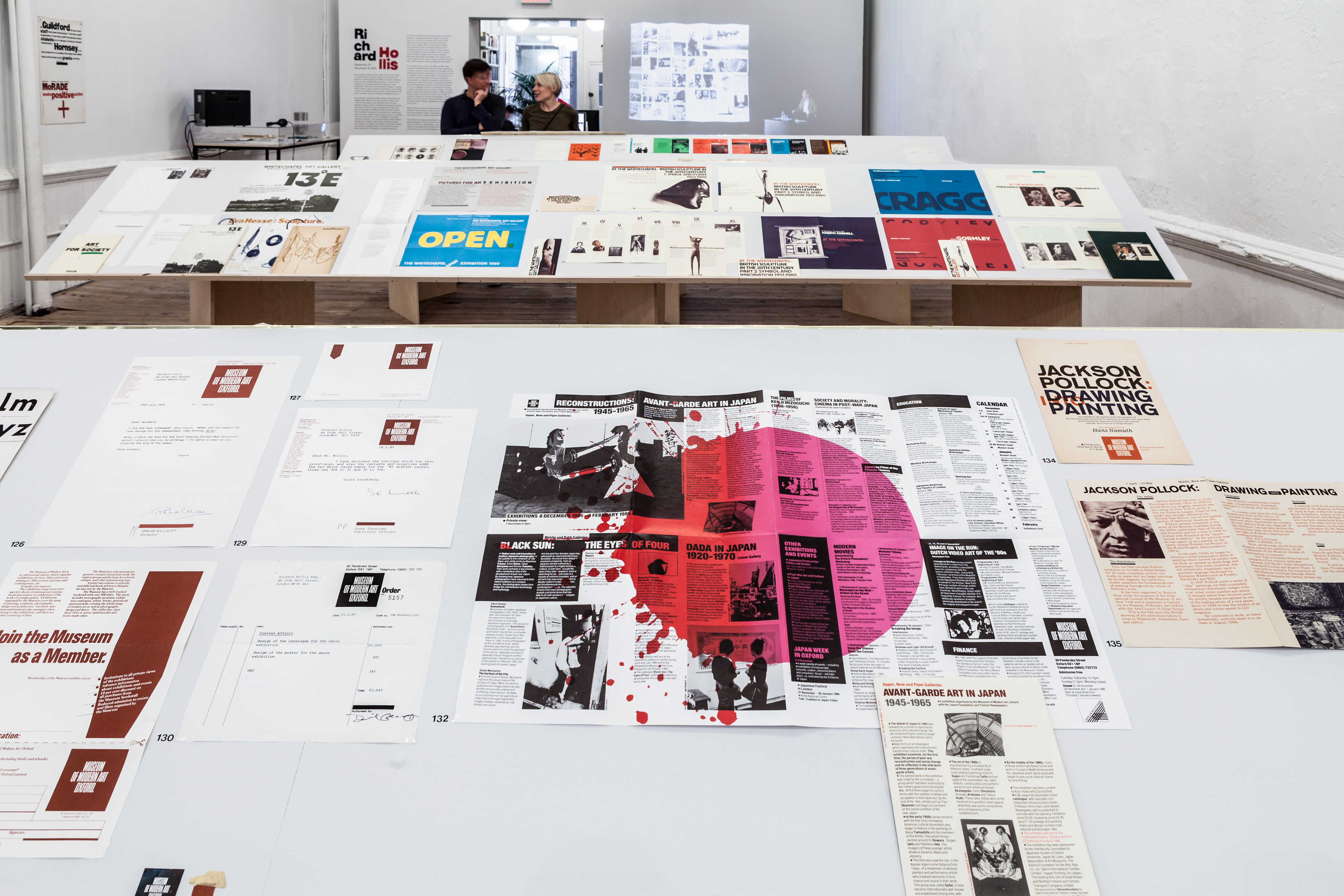 In the foreground, one side of a book stand displaying various posters and documents. Two more in the background include more items. Two people congregate in a coversation around a bookstand in the background.