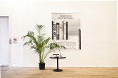 A large potted fern sits to the right of an elevator door in a light-filled gallery space. To the right of the fern is a low, round table with a pen and open booklet on top of it. A large black and white image installed on the gallery wall behind the fern shows several art books lined up next to one another on a blank surface. Text above the books reads, 