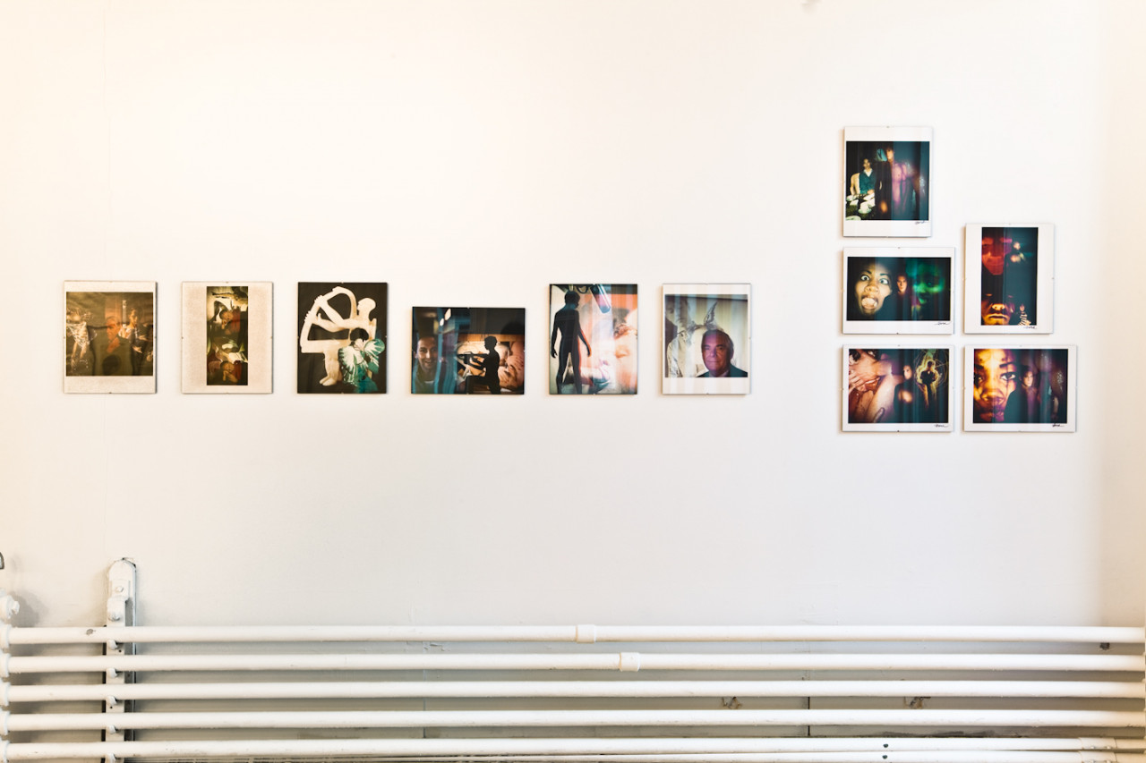 A series of photographs are installed on a white gallery wall above a series of pipes that run horizontally along the wall. The photographs depict figures superimposed by saturated imagery.