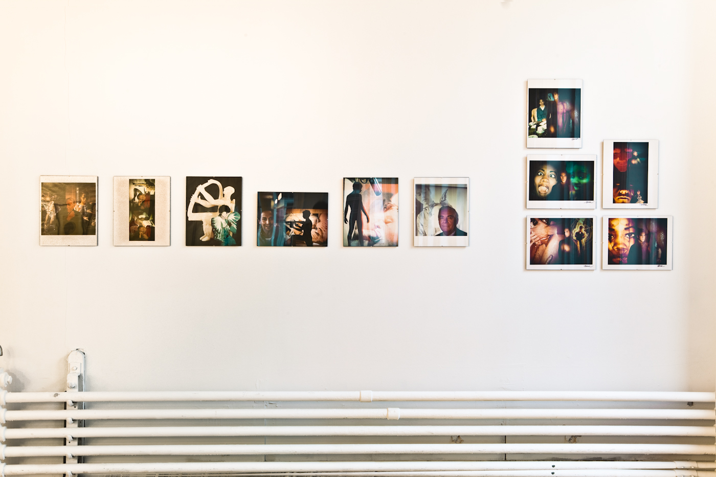 A series of photographs are installed on a white gallery wall above a series of pipes that run horizontally along the wall. The photographs depict figures superimposed by saturated imagery.