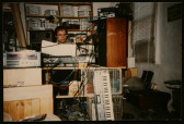 A vintage photograph of a man wearing black headphones, sitting behind a table in a cluttered room full of music equipment. A keyboard leans against the table, which is covered in AV equipment and snaking wires.