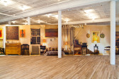 A large photo decal runs along the entire length of a long, horizontal wall in a light-filled gallery space with wooden floors. The photo decal depicts a to-scale, warmly-lit domestic space. A wooden chest of drawers, radiators, various shelves, and a low bed covered in a quilt fill the space. A dart board hangs on the wall, along with various paintings.