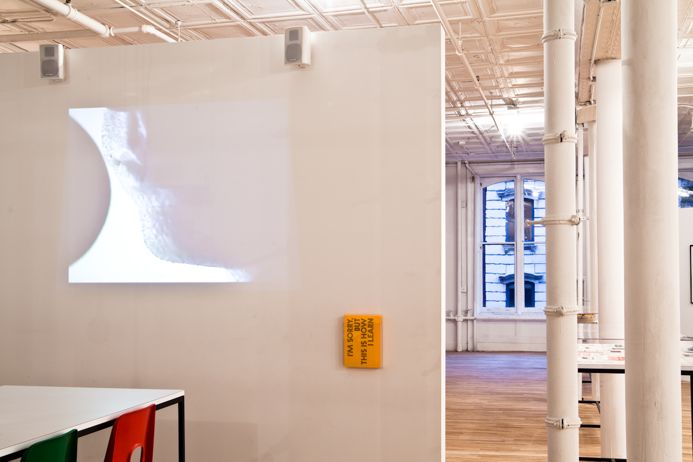 A projection of a close-up image of an unidentified figure's mouth and chin is displayed on a wall of a light-filled gallery space. Two small speakers sit above the projection at its top right and left corners. A series of yellow cards sit in a clear container mounted on the wall to the right of the projector. Bold text printed on the front of the cards reads: 