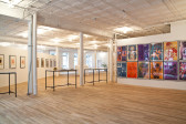 Three vitrines are displayed in a light-filled gallery space with light wooden floors. The vitrines sit in between three white columns to the image's left side. On the right, a series of large, colorful prints featuring graphic, abstracted letters overlaid over various abstract shapes, patterns, and printed figures are displayed on a gallery wall.