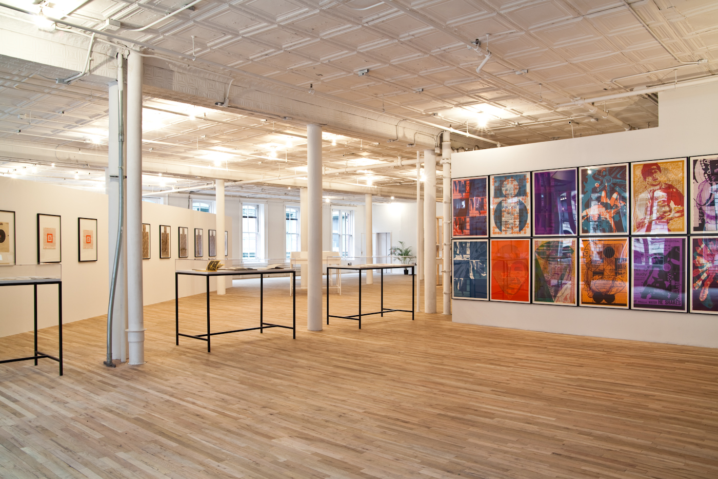 Three vitrines are displayed in a light-filled gallery space with light wooden floors. The vitrines sit in between three white columns to the image's left side. On the right, a series of large, colorful prints featuring graphic, abstracted letters overlaid over various abstract shapes, patterns, and printed figures are displayed on a gallery wall.