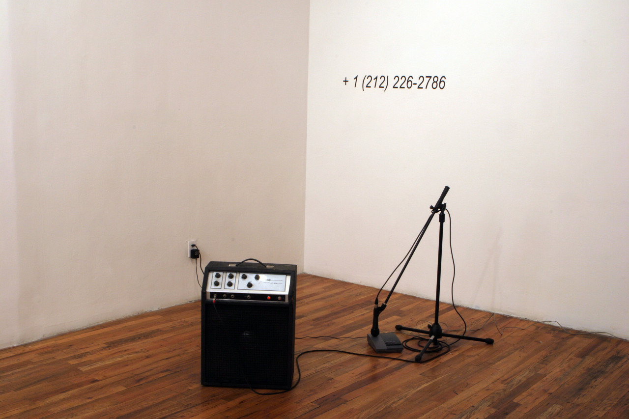 A microphone connected to a stand and an amp sit on the floor of a gallery space. The microphone stand has been adjusted such that the microphone is pointed down and hovers directly over a small answering machine on the ground. Wall vinyl installed on the wall directly behind the microphone stand displays a phone number: "+1 (212) 226-2786)."