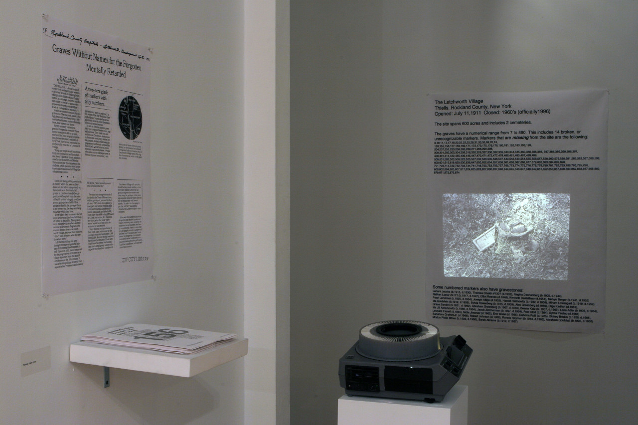 A slide projector sits in a dimly lit corner of a gallery space. The device projects an image of rocks and other debris onto a poster hung on the wall, which lists various statistics related to The Letchworth Village, a residential commuinty in Rockland County, New York. A second poster hangs on a wall to the left of the slide projector. A stack of papers rests on a small support below the poster.