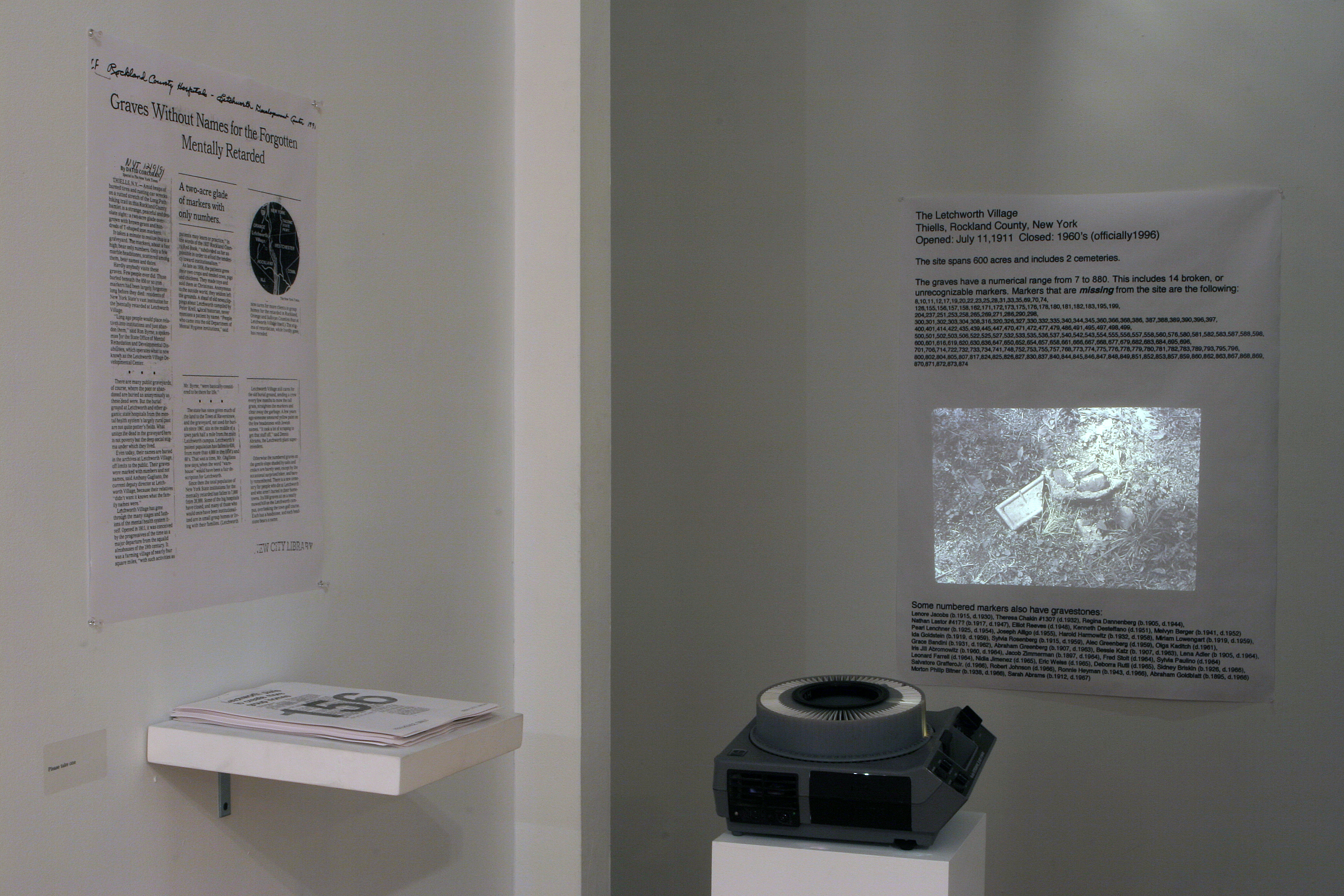 A slide projector sits in a dimly lit corner of a gallery space. The device projects an image of rocks and other debris onto a poster hung on the wall, which lists various statistics related to The Letchworth Village, a residential commuinty in Rockland County, New York. A second poster hangs on a wall to the left of the slide projector. A stack of papers rests on a small support below the poster.