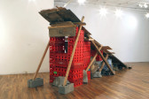 Bright red milk crates arranged on top of one another in descending height in a gallery space. Wooden planks are balanced on top of the crates and supported by longer wooden planks that rest against cinderblocks along the side of the structure.