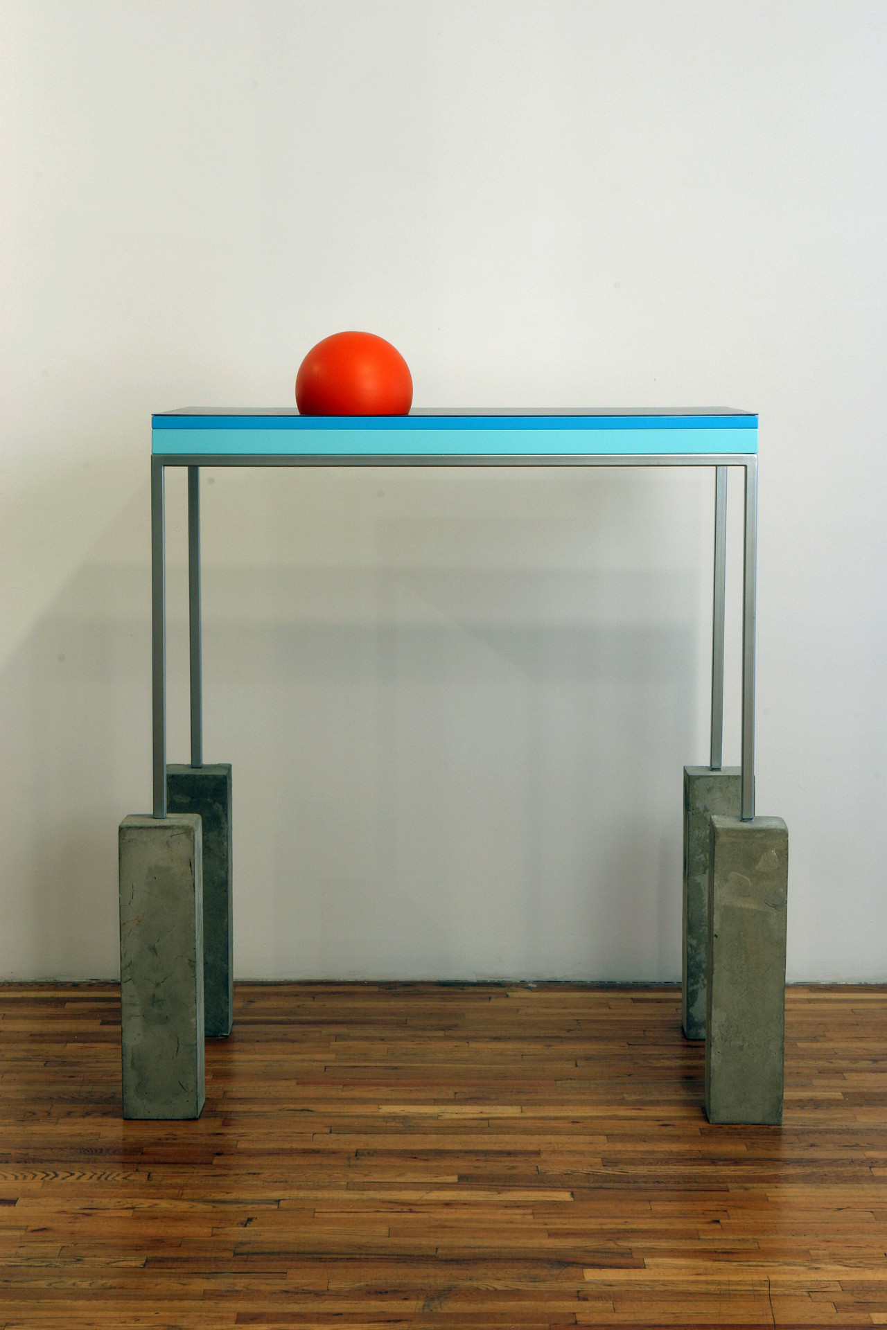 A metal desk with each of its legs perched on top of four vertical cinderblocks. The side of the desk is decorated with a dark blue and aqua blue stripe. A bright red ball rests on top of the desk.