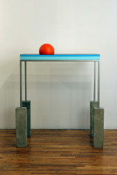 A metal desk with each of its legs perched on top of four vertical cinderblocks. The side of the desk is decorated with a dark blue and aqua blue stripe. A bright red ball rests on top of the desk.