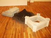 Two white models of large buildings sitting on a wooden floor. Between the two models is a large mound of dirt.