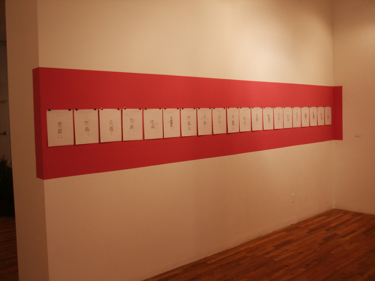 A red stripe is painted on a white wall. Hung in the red stripe are twenty sheets of white paper with a single figure drawn on each one.