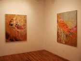 Two paintings hung on white walls meeting in a corner. The paintings are colorful, predominantly pink and red.