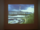 An image of a factory with smokestacks on concrete, just behind a body of water surrounded by long grasses.