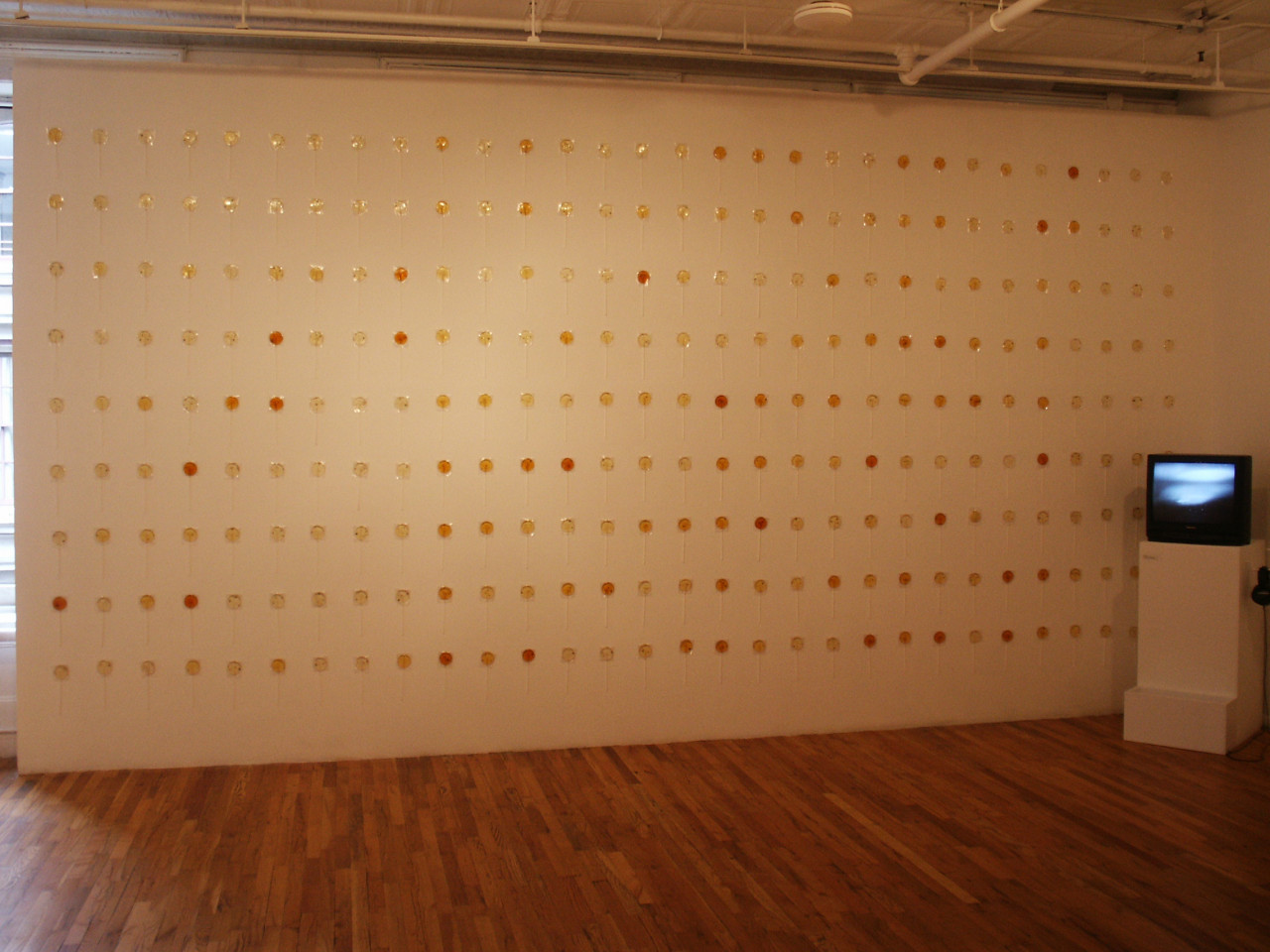A white wall with orange and yellow lollipops arranged in lines. In front of the wall is a small television on a white plinth.