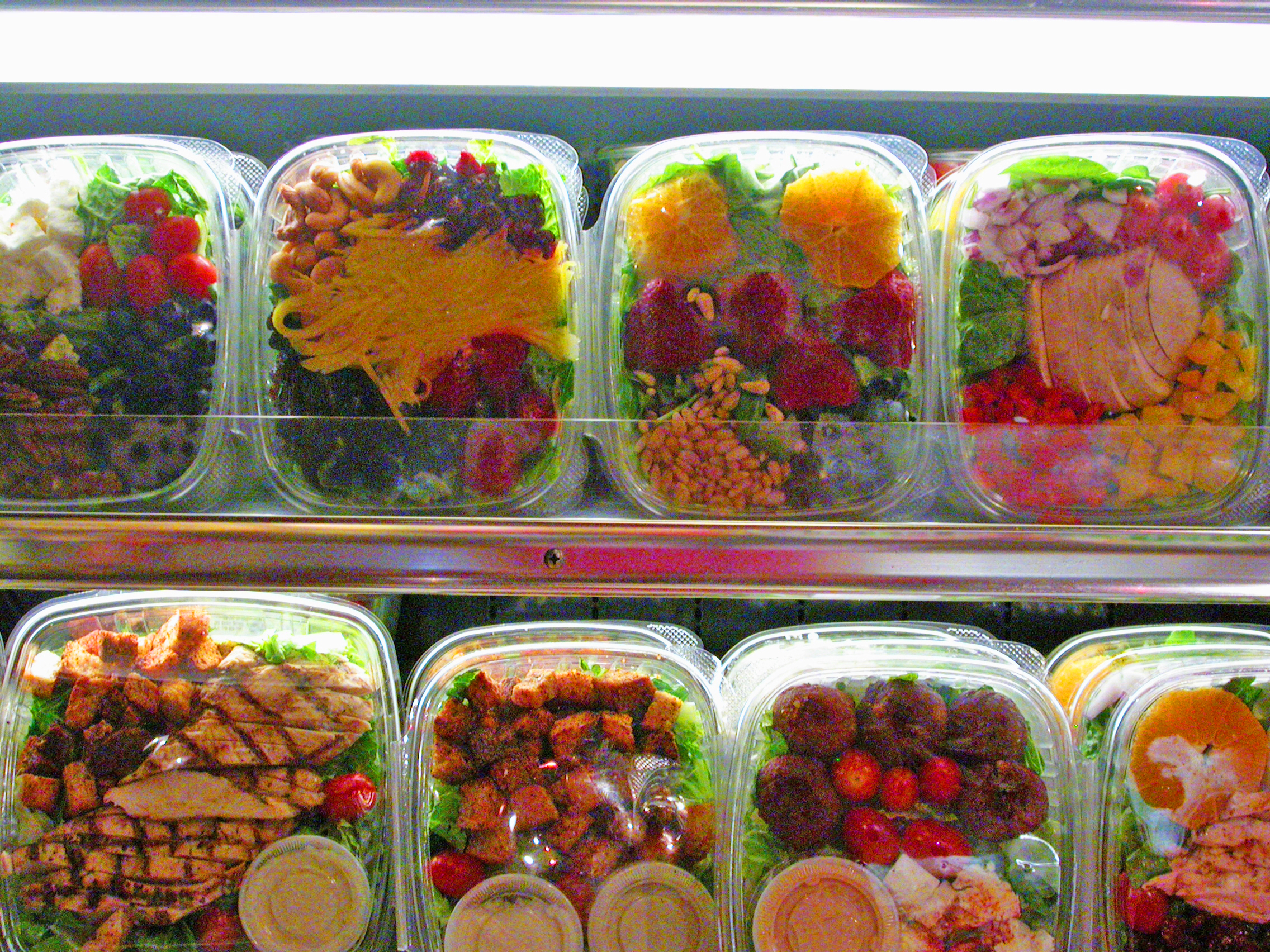 Close up of two rows of pre-packaged salads in plastic containers on the shelves of a grocery store refrigerator.