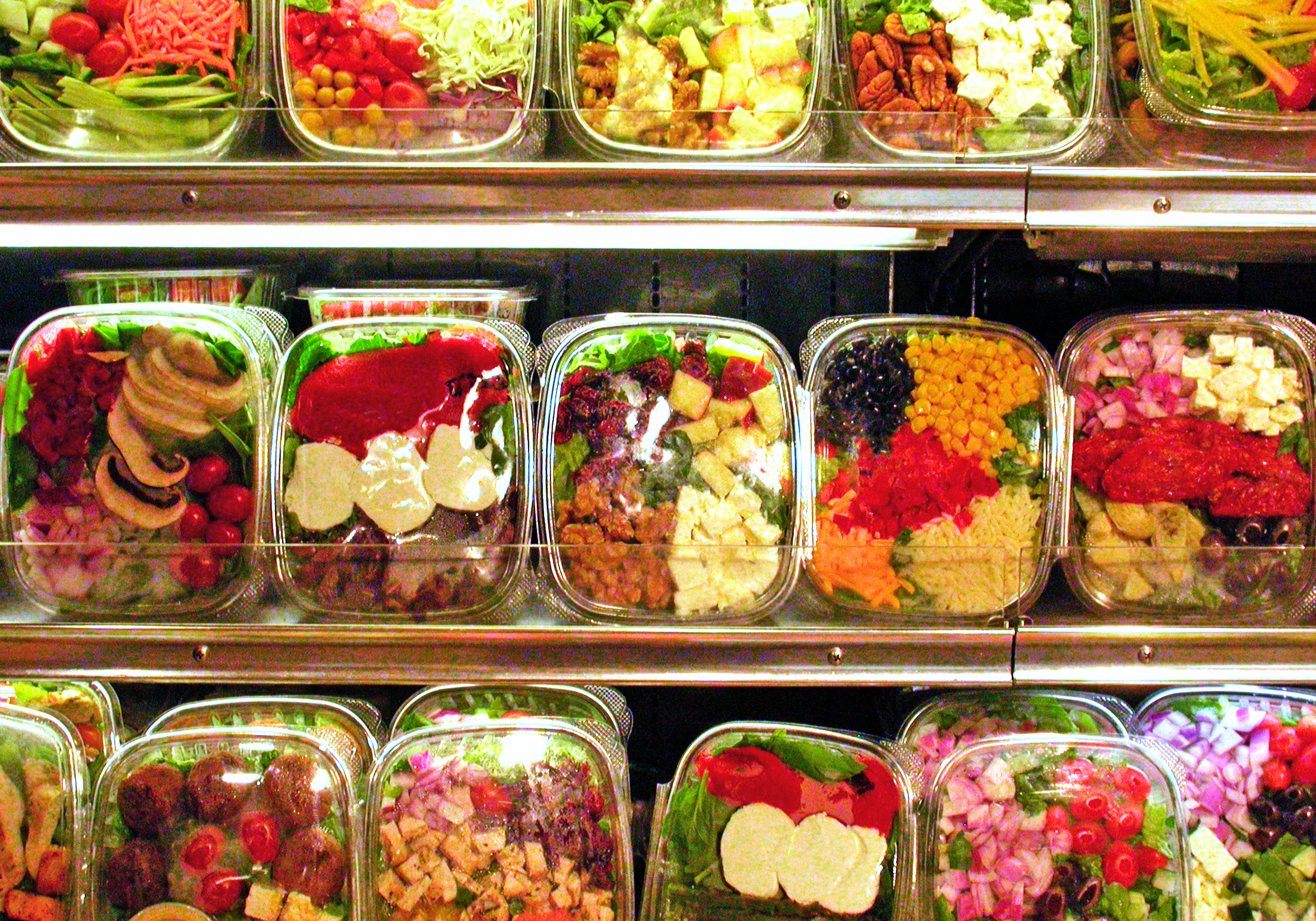 Close up of three rows of pre-packaged salads in plastic containers on the shelves of a grocery store refrigerator.