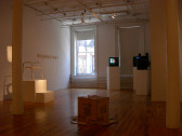 Several artworks sit in a dimly lit gallery space, illuminated by light from an open gallery window. Three video monitors are installed on plinths to the left of the image. In front of them and closest to the viewer, an overturned cardboard box rests on the floor. To the left, a stool-like structure sits on a plinth illuminated by a bright overhead light. Behind this structure, a series of small works on paper are displayed on a gallery wall.