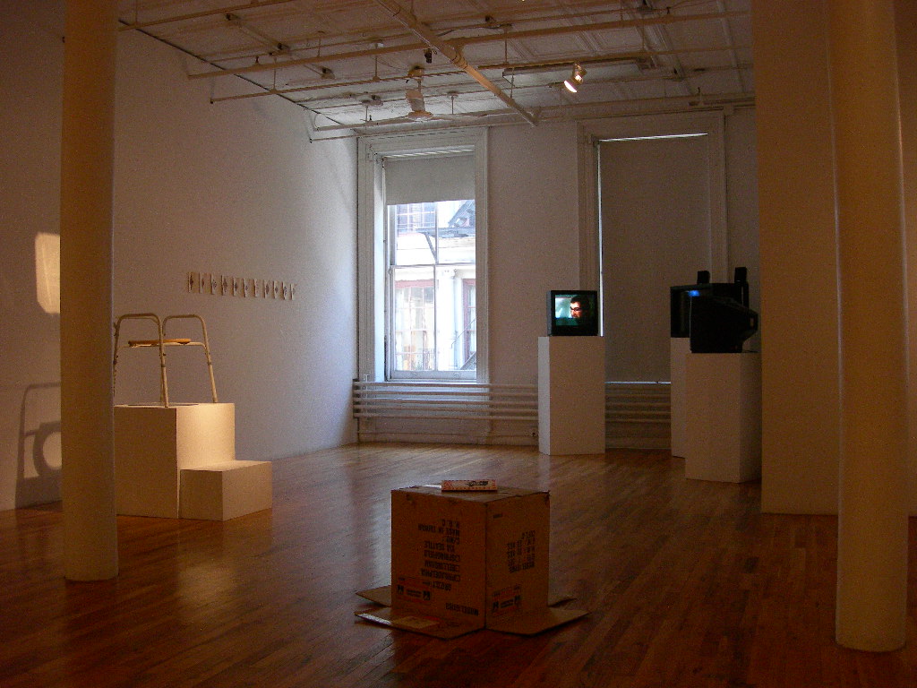 Several artworks sit in a dimly lit gallery space, illuminated by light from an open gallery window. Three video monitors are installed on plinths to the left of the image. In front of them and closest to the viewer, an overturned cardboard box rests on the floor. To the left, a stool-like structure sits on a plinth illuminated by a bright overhead light. Behind this structure, a series of small works on paper are displayed on a gallery wall.