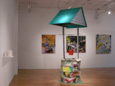 A large structure resembling a well is displayed in a gallery space with wooden floors. The sides of the well are constructed from spray-painted cinder blocks. Supported above the well's opening and a hanging, red bucket is a structure resembling the top portion of a house roof. Behind the well, three large paintings composed of colorful, gestural marks are installed on the gallery wall.