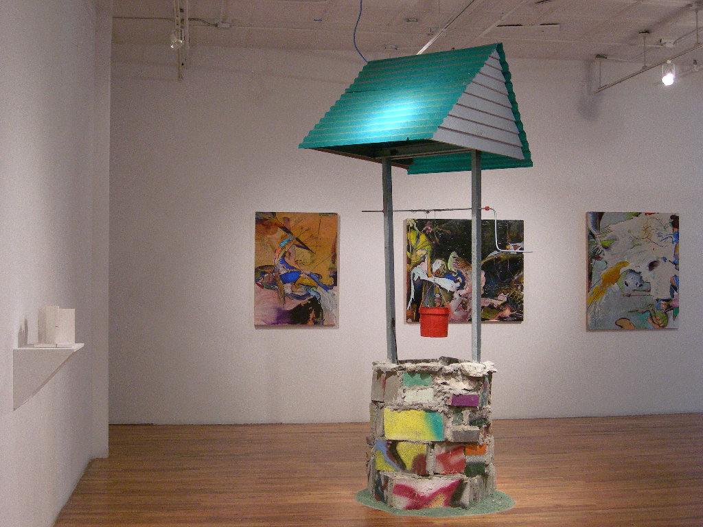 A large structure resembling a well is displayed in a gallery space with wooden floors. The sides of the well are constructed from spray-painted cinder blocks. Supported above the well