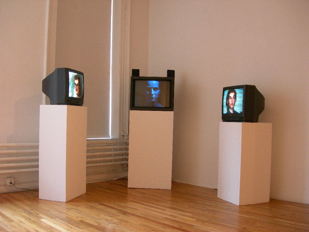 Three black television monitors installed on white plinths in a gallery space in a semi-circluar formation. Faces of three disparate figures are displayed on each screen. The face displayed in the center monitor