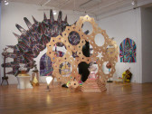 Several geometric structures of varying sizes are displayed in close, overlapping clusters in a gallery space. The largest structure of the group nearly touches the gallery ceiling is composed of an array of flat pentagons that form circular, symmetrical patterns. Behind this structure is a large archway-shaped wall decal showing further colorful arrays and patterns. Several other, smaller pieces with similar geometric forms cluster on the floor around these structures.
