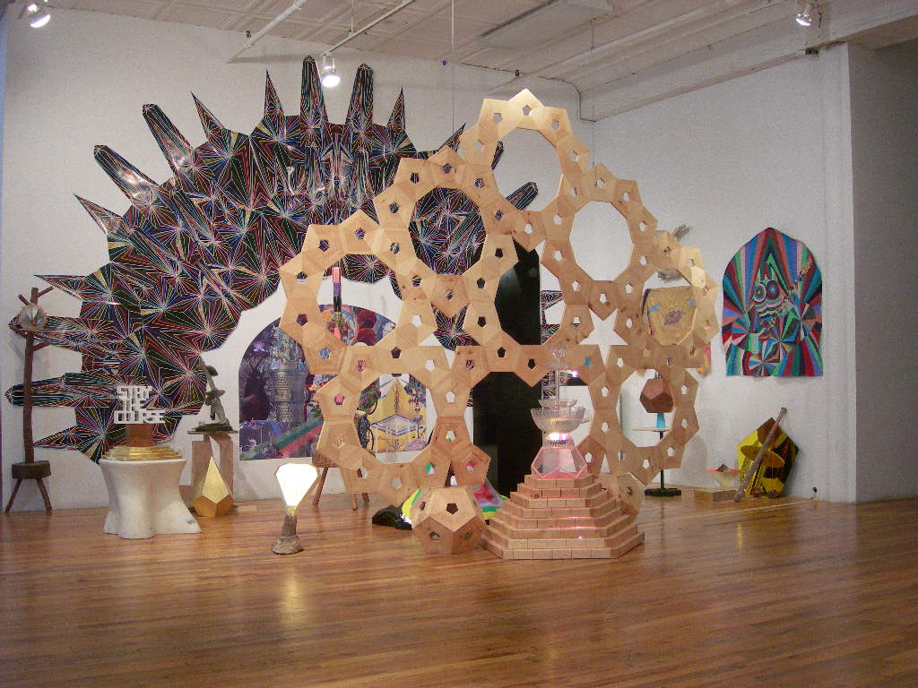 Several geometric structures of varying sizes are displayed in close, overlapping clusters in a gallery space. The largest structure of the group nearly touches the gallery ceiling is composed of an array of flat pentagons that form circular, symmetrical patterns. Behind this structure is a large archway-shaped wall decal showing further colorful arrays and patterns. Several other, smaller pieces with similar geometric forms cluster on the floor around these structures.