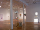 Several sculptures and 2D works sit in a large gallery space with wooden floors and white columns. To the right, a stool-like object sits on a white plinth, illuminated by a bright overhead light. On the ground in the center of the image, an overturned cardboard box rests on the floor. A colorful structure resembling a well is installed in the background.