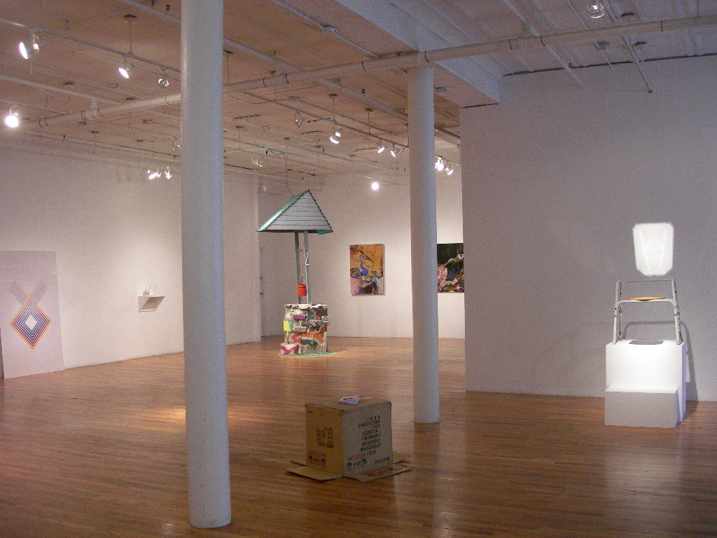 Several sculptures and 2D works sit in a large gallery space with wooden floors and white columns. To the right, a stool-like object sits on a white plinth, illuminated by a bright overhead light. On the ground in the center of the image, an overturned cardboard box rests on the floor. A colorful structure resembling a well is installed in the background.