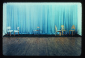A color photograph of six chairs in front of a blue curtain.