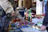 A person in army camouflage and a military gun grabs a handful of packaged cookies from a stand selling assorted candies, snacks, sodas, and other food.