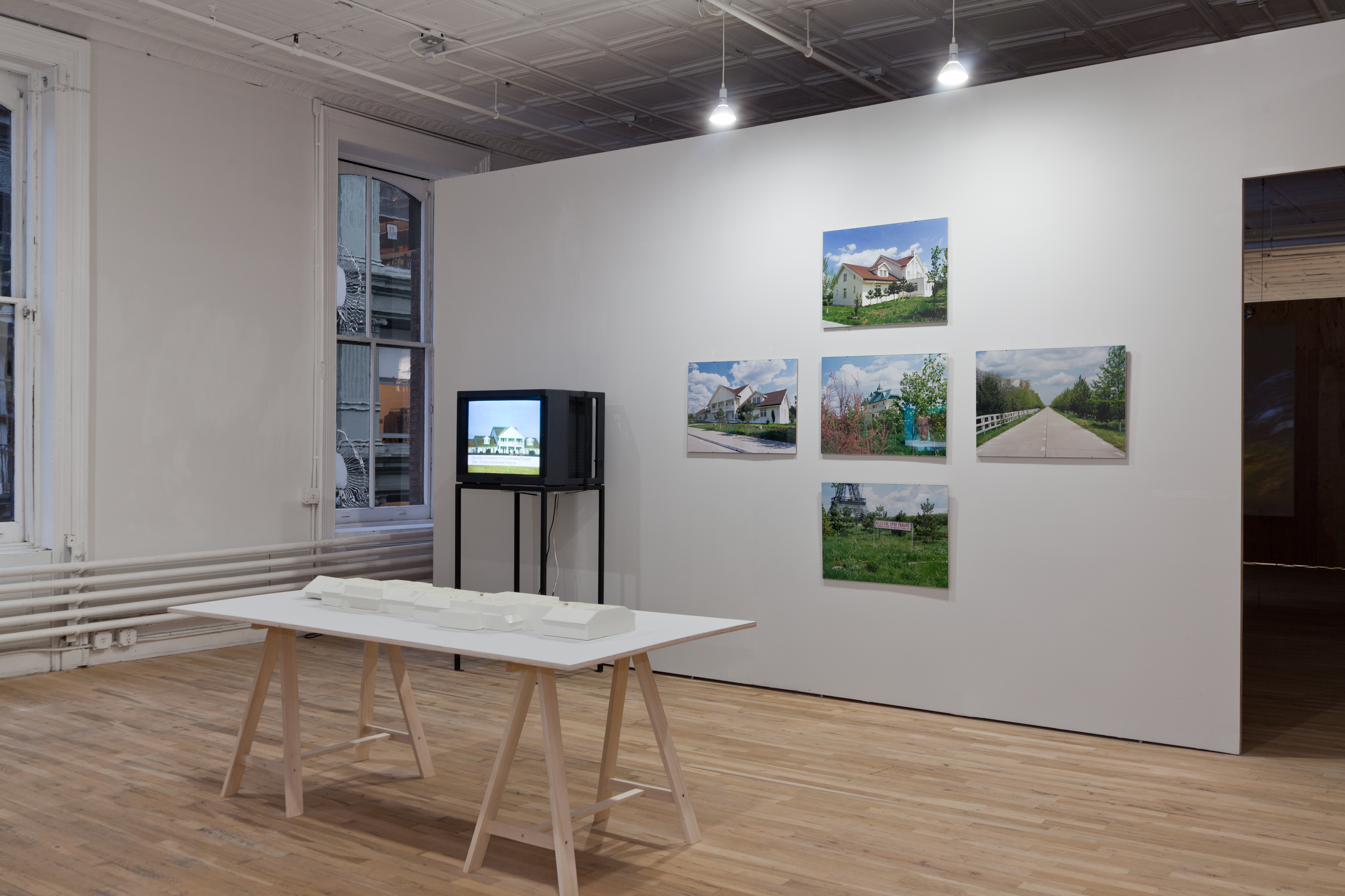 A white table holds a white model of a house. Behind it, a television on a stand displays an image of a large, white house. There is a white wall with five color photographs arranged in a cross formation with images of a large house and yard.