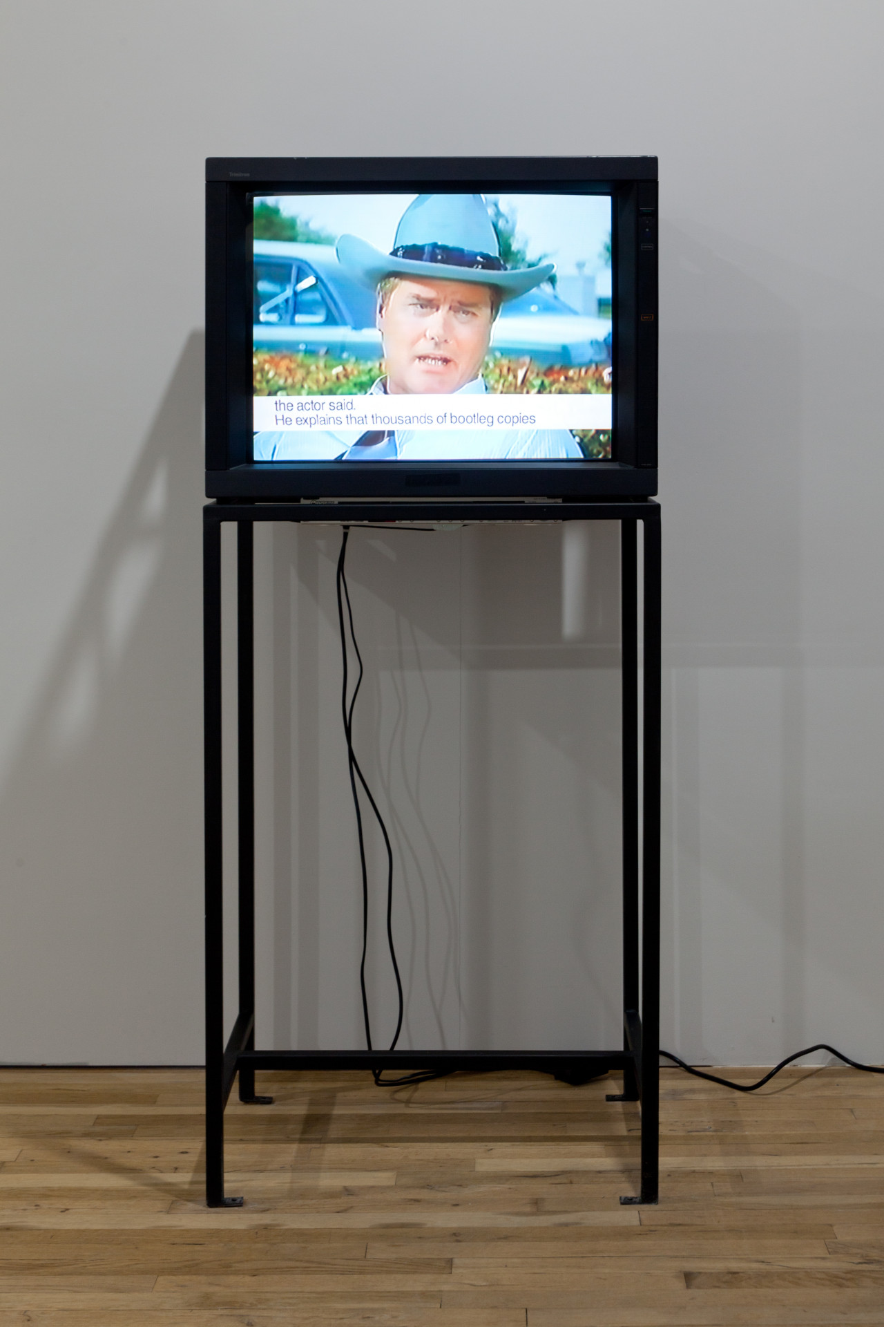A television on a metal stand shows an image of a man in a cowboy hat. A white bar with black text on the bottom of the screen reads, "The actor said. He explains that thousands of bootleg copies."