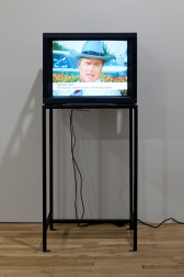 A television on a metal stand shows an image of a man in a cowboy hat. A white bar with black text on the bottom of the screen reads, 