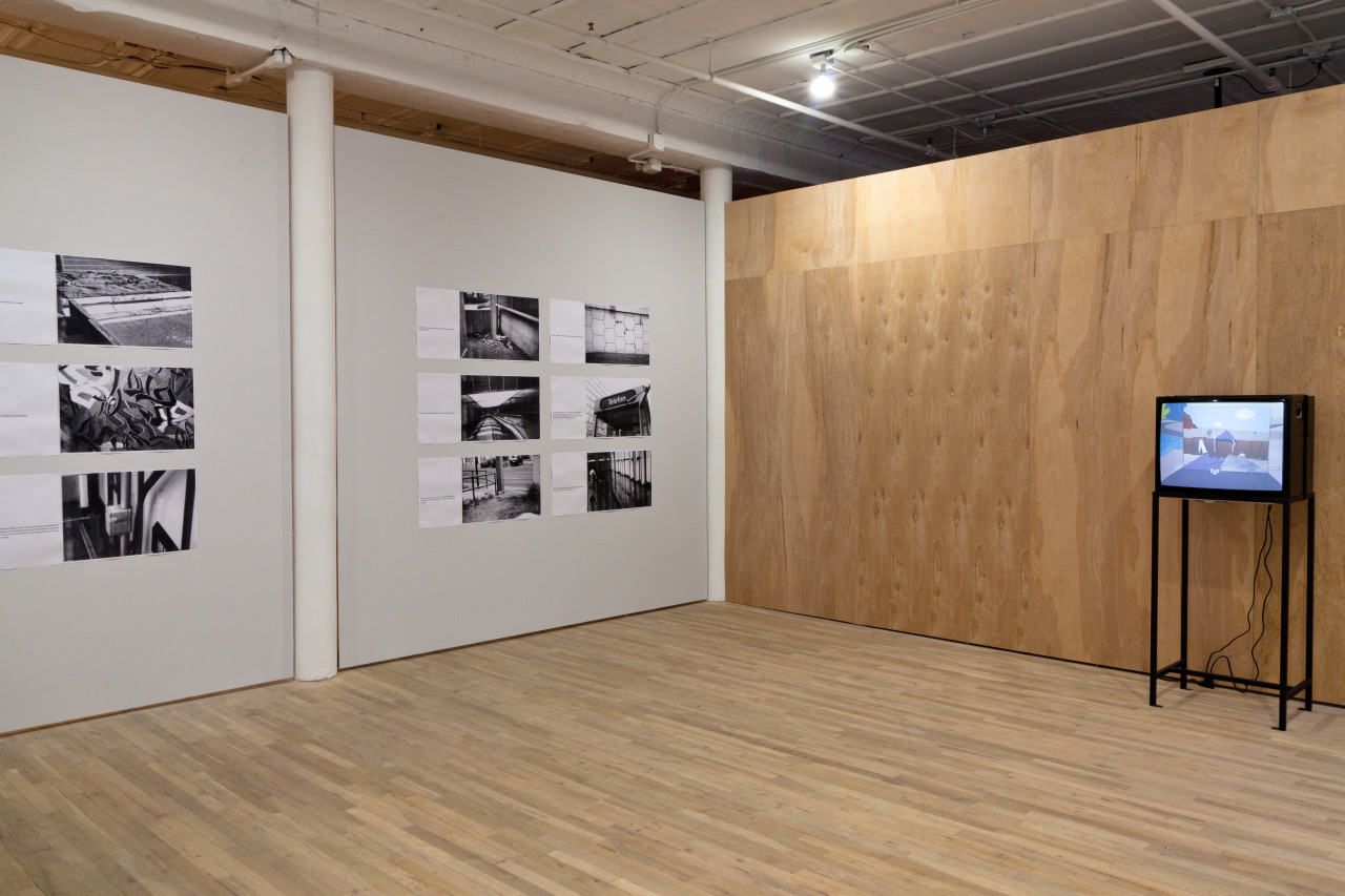 An installation shot of two gallery walls, one white and one unpainted wood, meeting in a corner. The white wall displays nine images with accompanying text. The wood wall has an old television on a metal stand, showing a cartoon image.