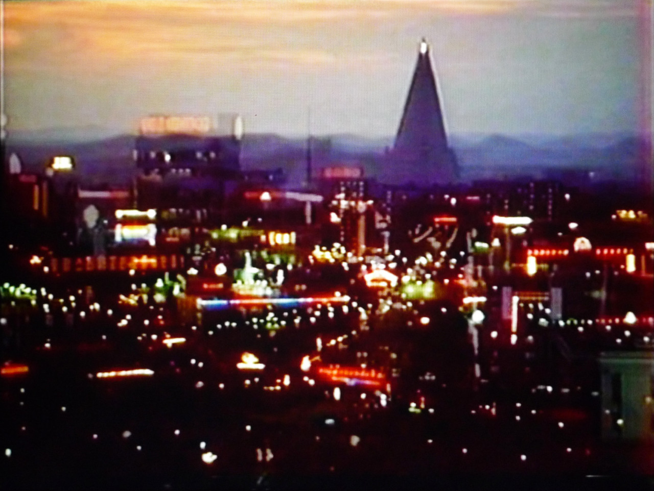 A slightly blurry image of a city in the evening. An outline of a tall, triangular building is visible against a sunset sky.